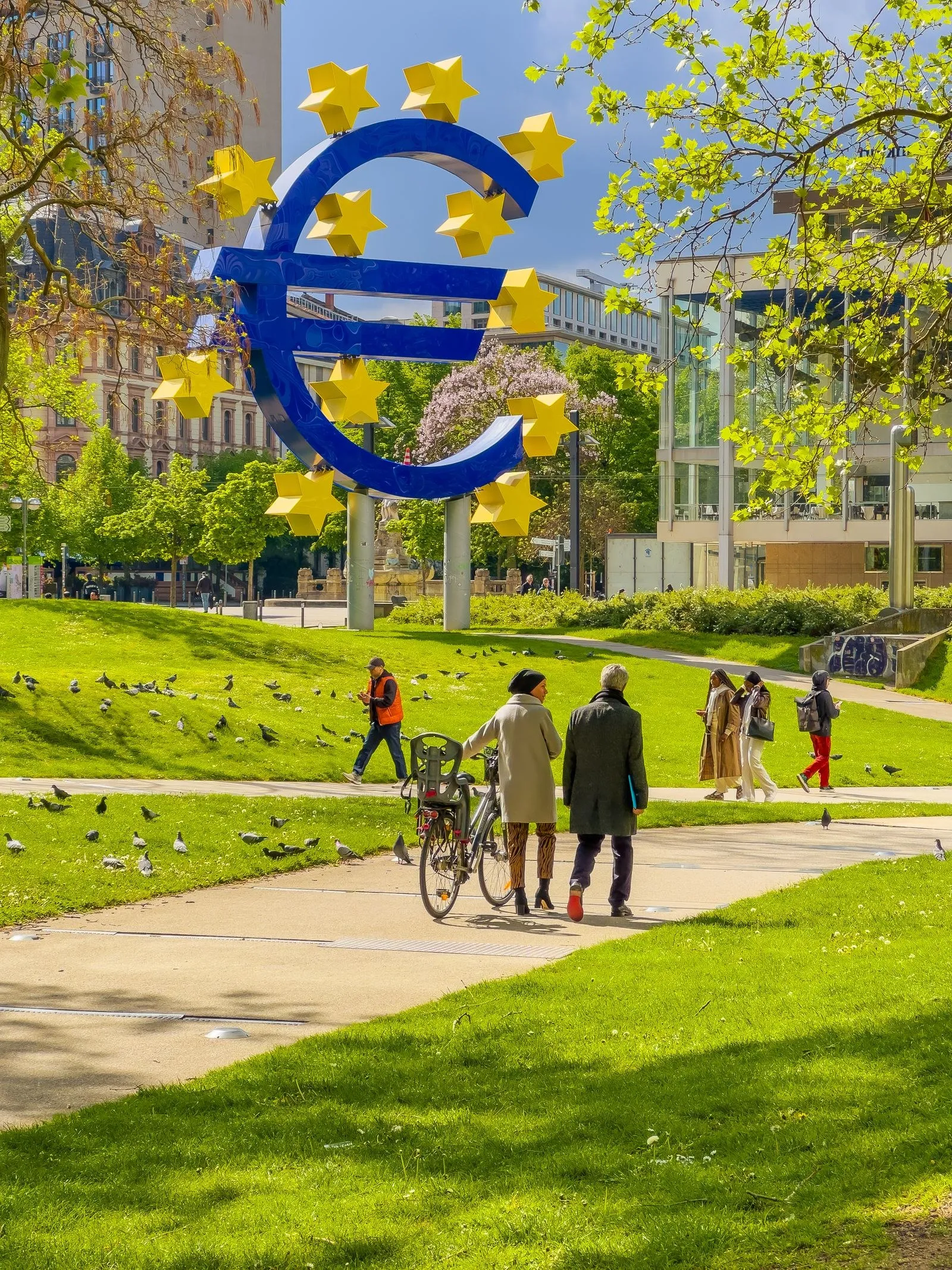 The Euro sculpture in Frankfurt, a large blue euro sign ringed by yellow stars, with people walking in the park around it