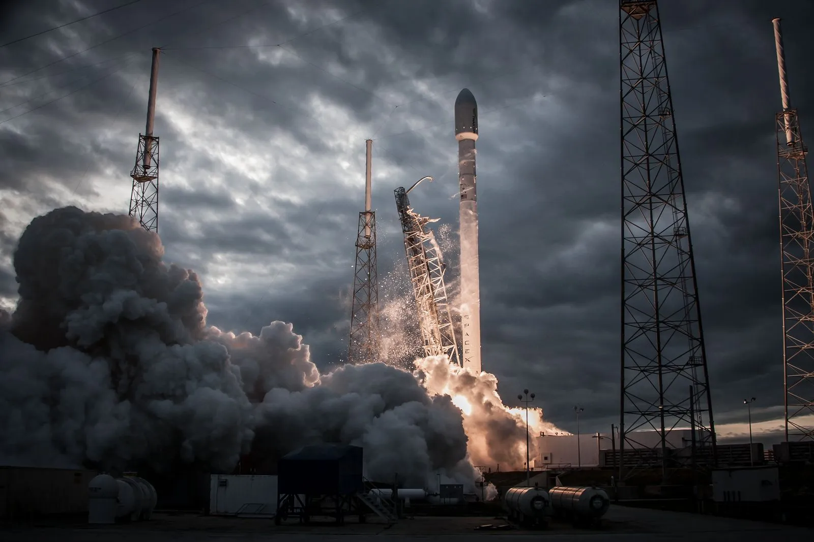 A SpaceX Falcon 9 rocket lifting off from the launch pad under an overcast sky, engulfed in smoke